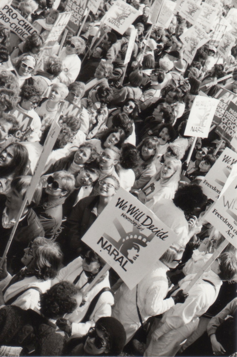 A black and white photograph taken from an angled overhead shows a large crowd of people, some of whom are wearing white, holding signs that say, “We Will Decide NARAL.”