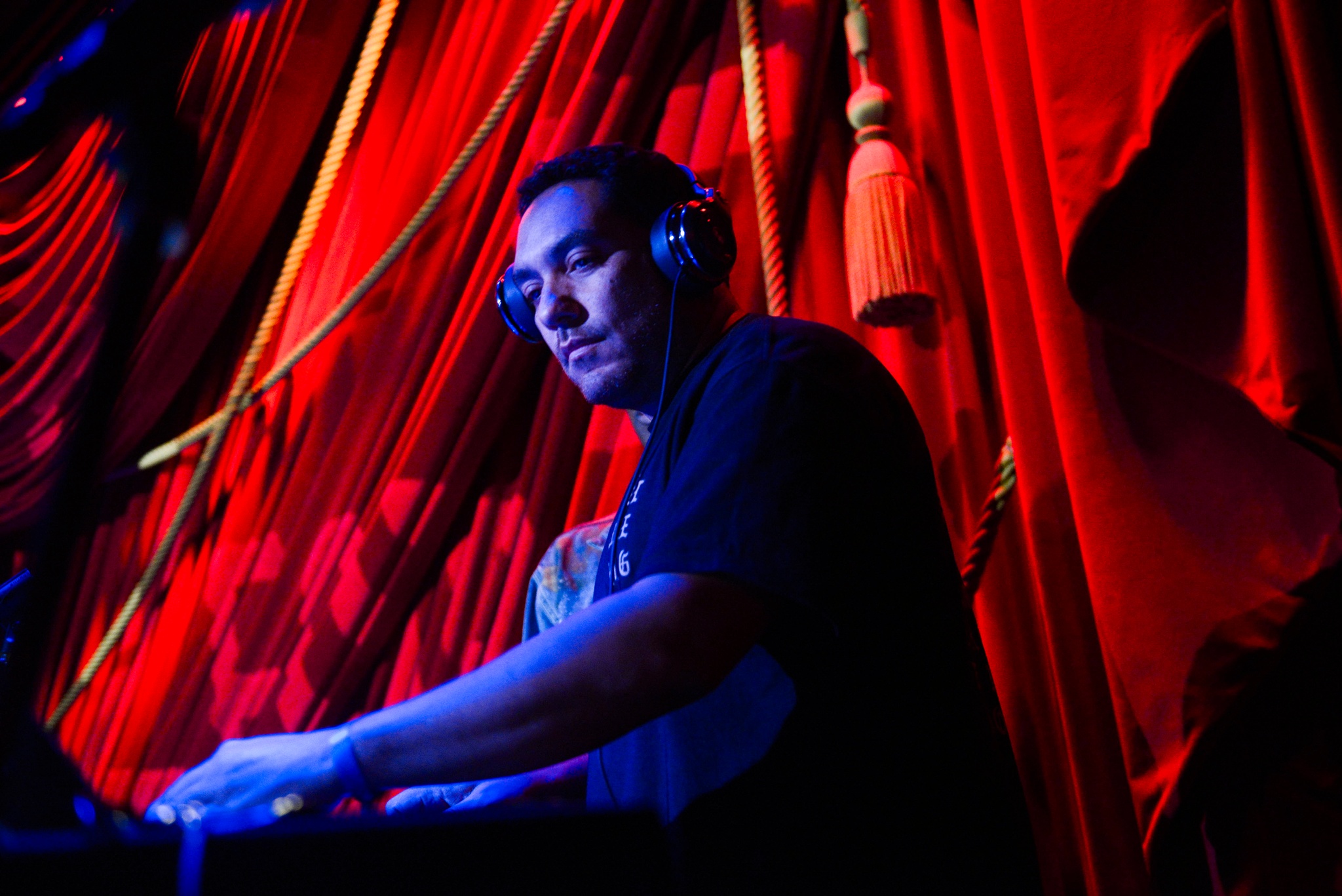 A DJ spinning bathed in blue light on his face and arms with a red curtain hanging behind him. He is seen from a low angle.