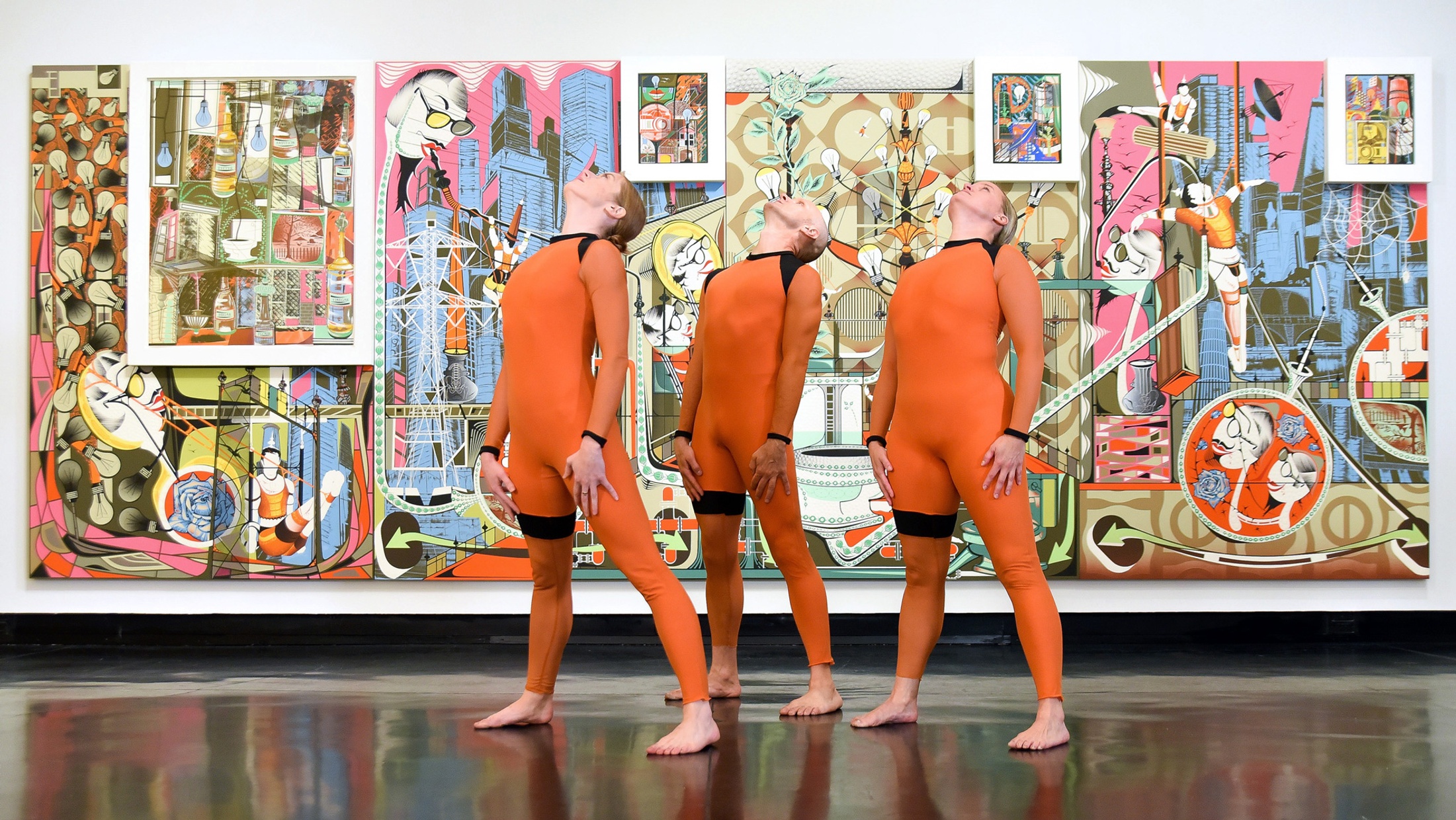 Three dancers in orange jumpsuits stand with their heads back in front of a large, abstract, colorful painting.