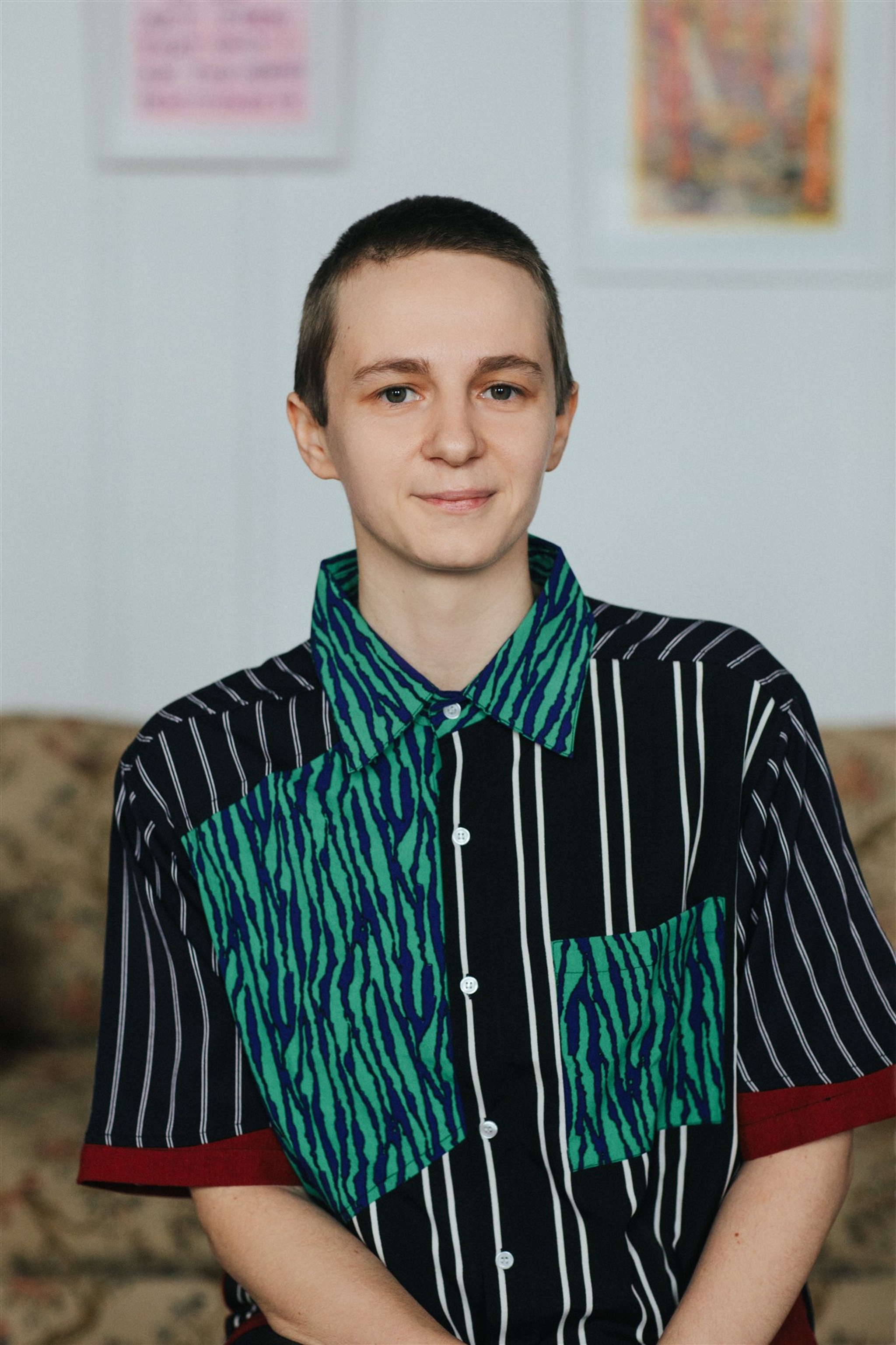 A portrait of Finnegan Shannon, a white person with short cropped hair who wears a multi-patterned green and blue short sleeve shirt. Behind them hang small artworks on a white wall.