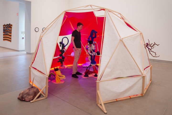 Geodesic tent with white fabric on the exterior and neon pink interior with uplighting, filled with bizarre alien flora and fauna sculptures.