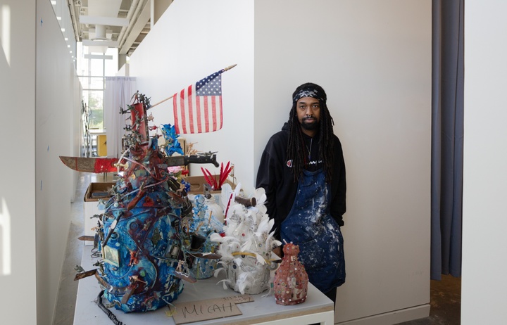 Micah Mickles leaning against a white wall with his sculpture on the left side