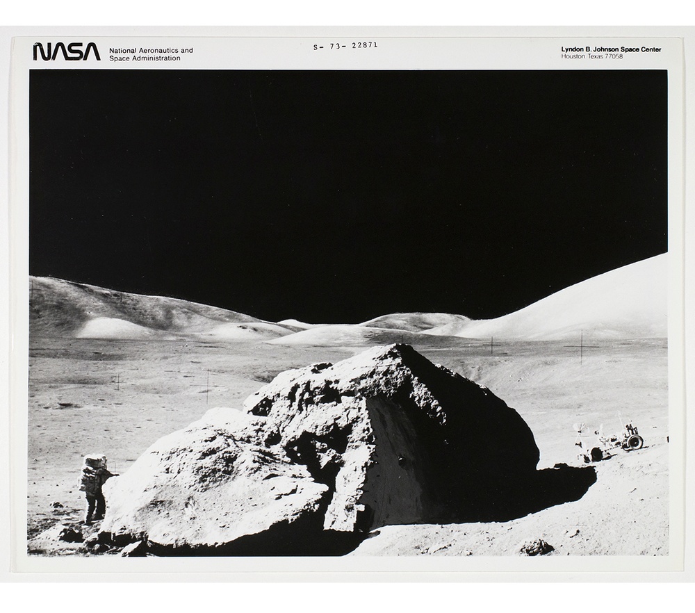 Black and white photograph of the moon’s surface, looks like a desert with a large rock and astronaut standing on one side the rock with a lunar roving vehicle on the other side.