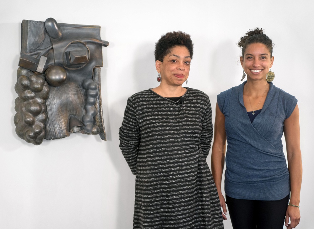 Syd Carpenter and Leah Penniman stand and smile for the camera with Syd's sculpture mounted on the wall behind them on the left side of the photo.