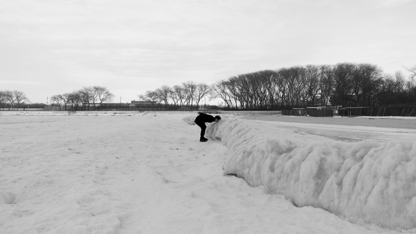 Black-and-white photo of an open, snowy landscape, with a person bent at the waist, their face close to a snowbank.