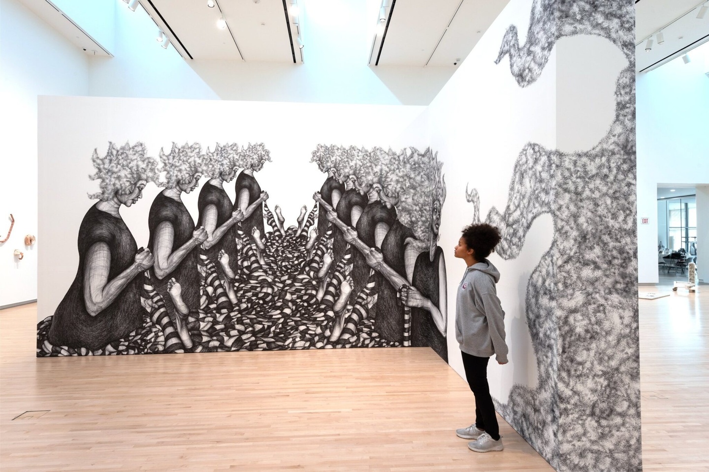 Inside a museum, a young Black woman stands next to a larger-than-life, black & white drawing installation composed on an L-shaped section of wall. On one wall, two rows of Black women remove striped socks from unseen figures' feet in an almost assembly line fashion. On the adjacent wall, a cloudlike formation of hair curls around the corner.