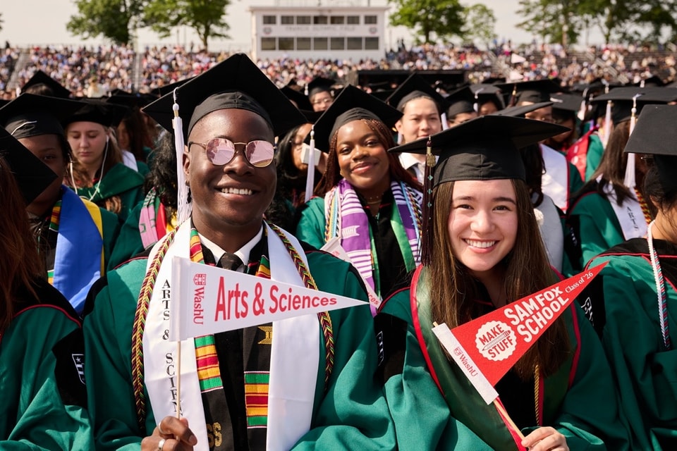 Two people wearing regalia and holding pennants stand next to each other and smile