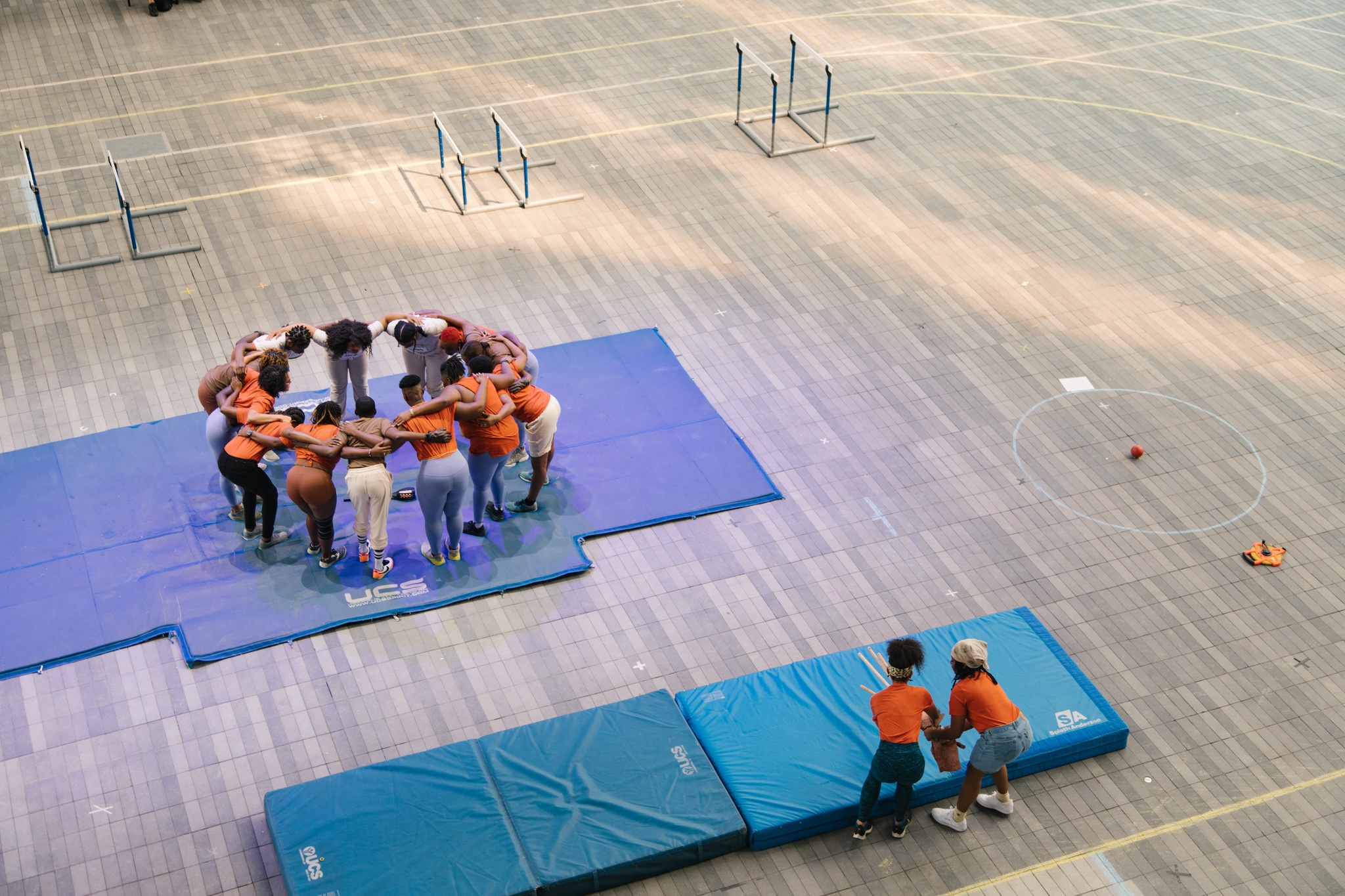 A group of track-and-field athletes huddle in a circle with arms interlaced around each other's shoulders. They stand on a blue atlhletic mat.