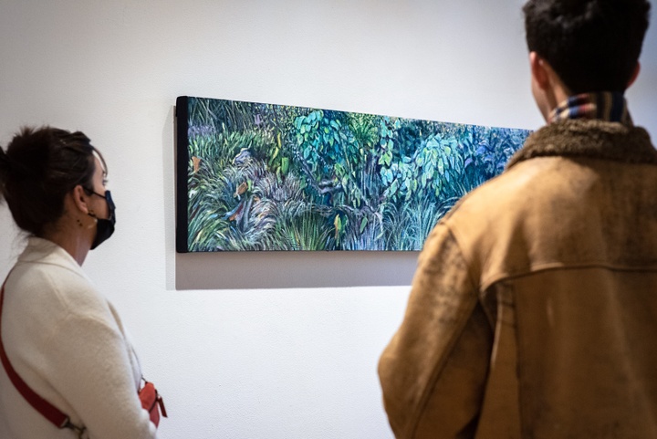 Two people look at a long, narrow painting of intricate blue and green tropical foliage.