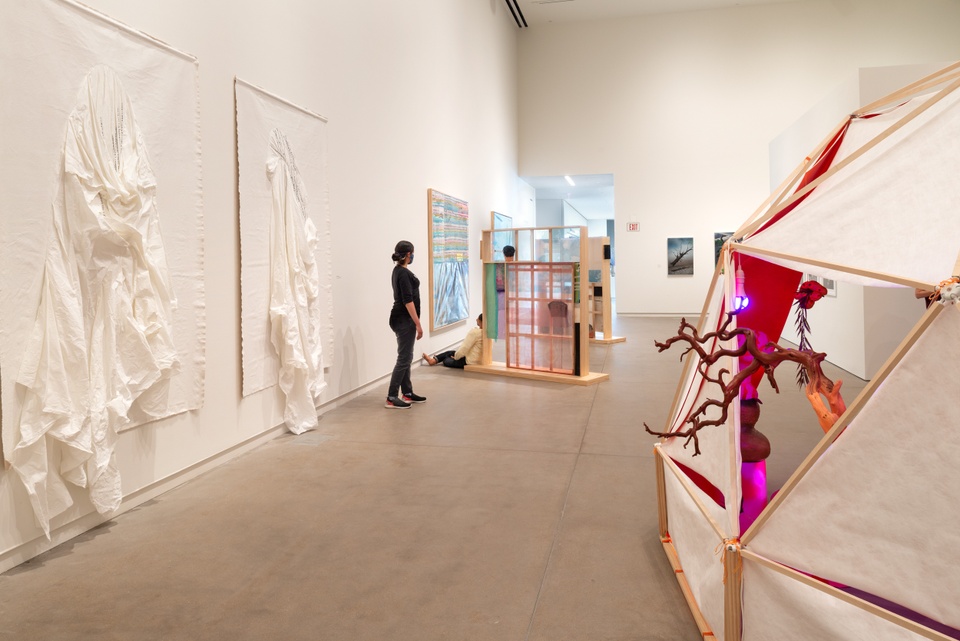 Overview shot of a gallery space with white fabric pieces on the wall and sculptural pieces on the floor.