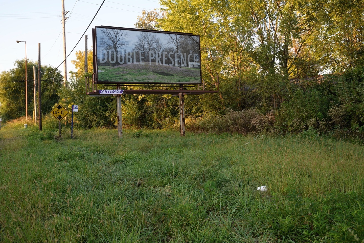 Billboard featuring a landscape with a mounded green hill and the words Double Presence in all-caps, set in an outdoor area with trees and grass.