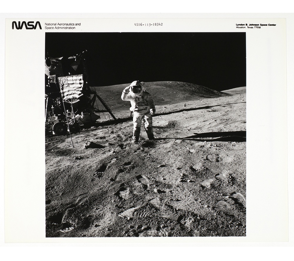 Black and white photograph of an astronaut on the moon saluting the American flag surrounded by foot prints with a lunar lander in the background.