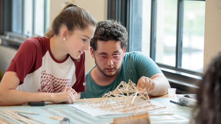 Two people inspect an architectural model built of wooden sticks.