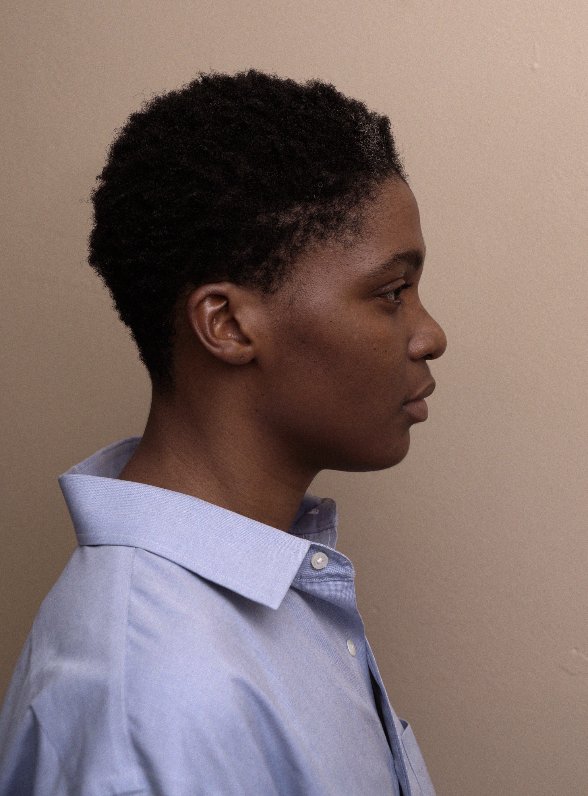 NIC Kay, a gender non-conforming (GNC) individual of Black descent, is prominently featured in this image. Their shortly cropped natural hair frames their face elegantly. NIC Kay’s profile is visible as they face to the left, radiating a quiet strength. They wear an oversized blue shirt that drapes loosely over their frame, standing out against the cream background.