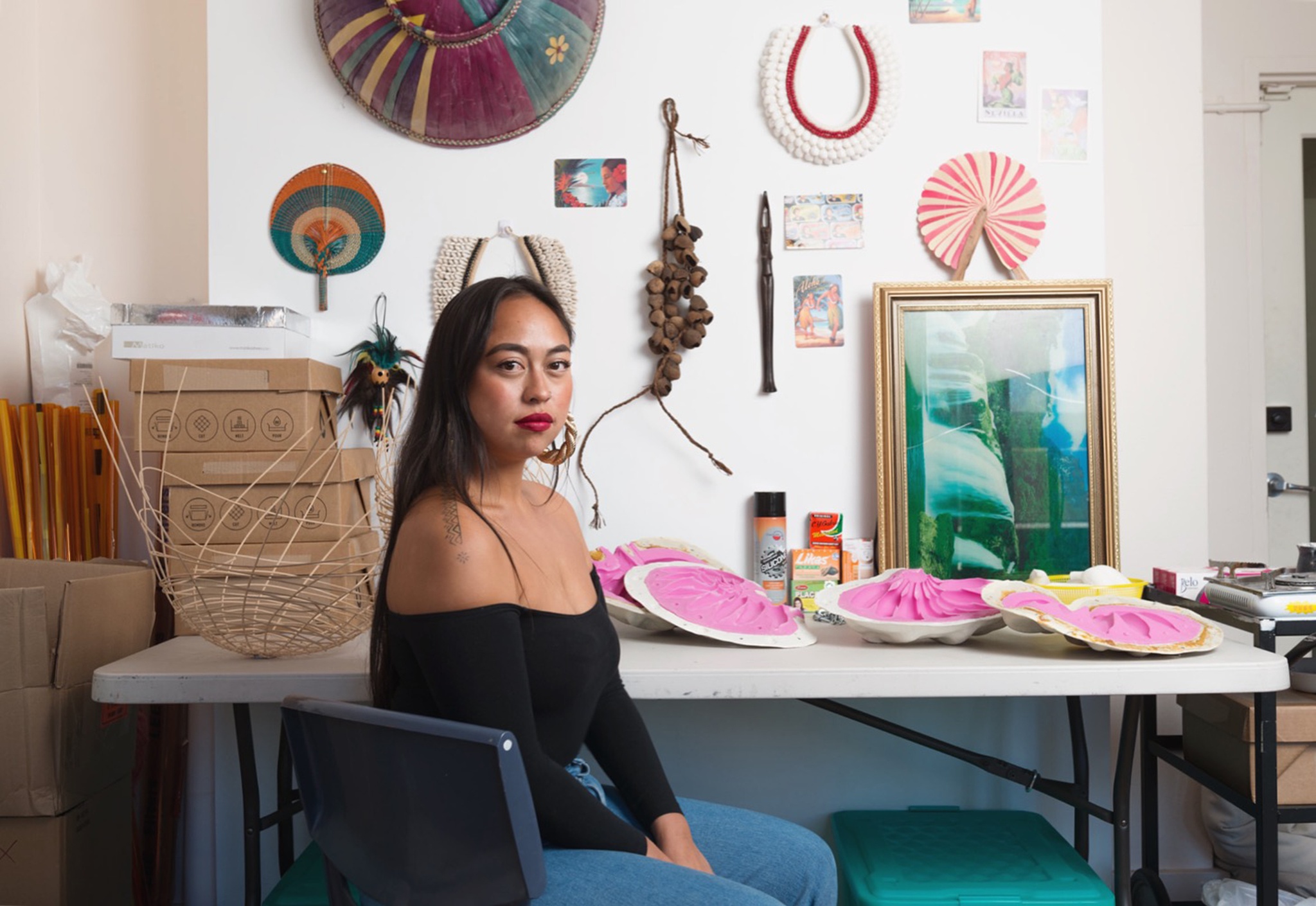 A photo of the artist Caroline Garcia sits in front of a table in her studio. On the wall behind her hang colorful fans, postcards, and other objects.