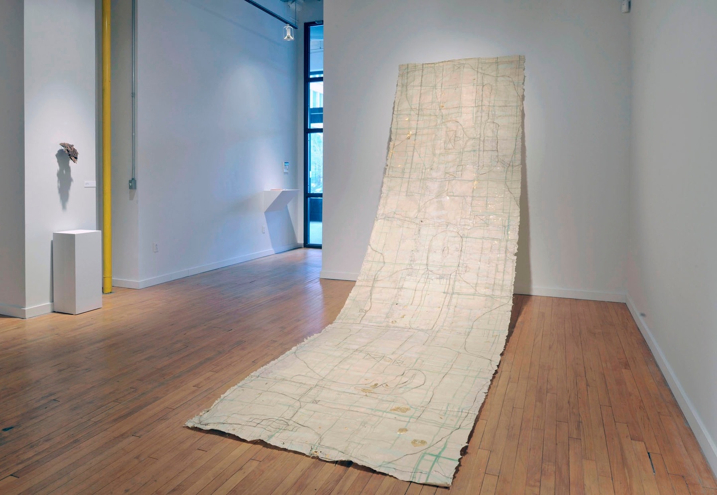 In a gallery space with white walls and light wooden floors, an installation: a rug/carpet hangs from the wall, extending to the foreground; it is white/cream-colored, its surface stitched/adorned with lines that thread across the length of the material.