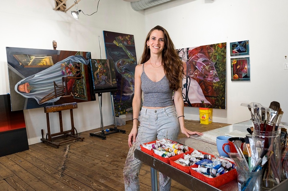 artist Maria Adesman standing in her studio next to a table with paint supplies and paintings on easels and hanging on the wall behind her
