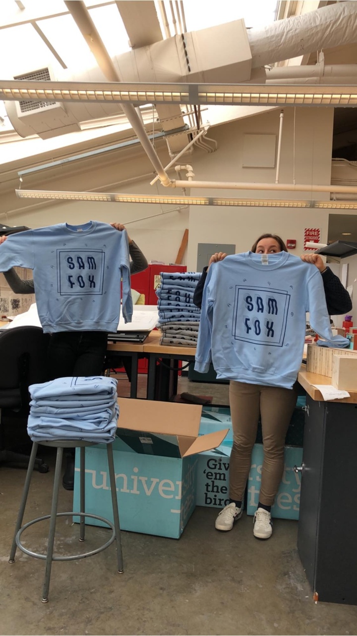 Two people hold up light blue shirts reading "Sam Fox" in front of their faces.