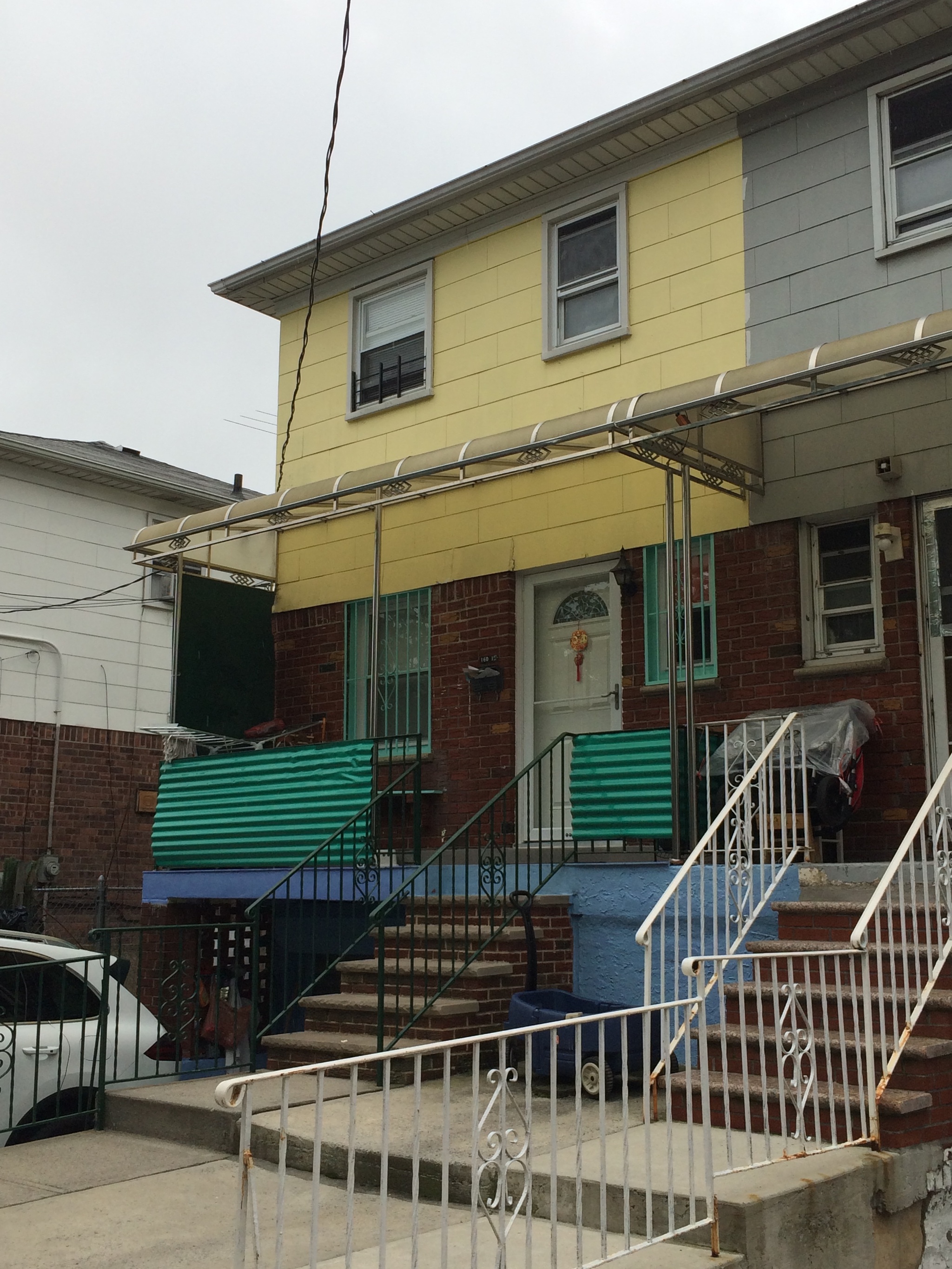 A rowhouse with yellow siding sits adjacent to an attached house painted gray. The yellow house is three stories including a basement level and the elevated front porch has plastic corrugated sheets for a railing. Both houses have wrought iron railings along their front stairways and sidewalks. 