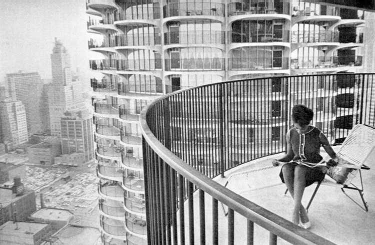 Black and white photo of Geraldine Johnson sitting on her Marina City balcony in Chicago; the balcony has a curved shape; in the background is the city skyline, including a prominent multistory building.