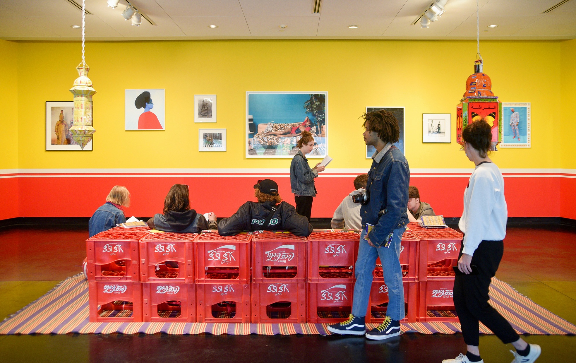 Two people walk past a group of people sitting in a couch made of Coca Cola crates looking at a yellow wall with artwork hung on it.