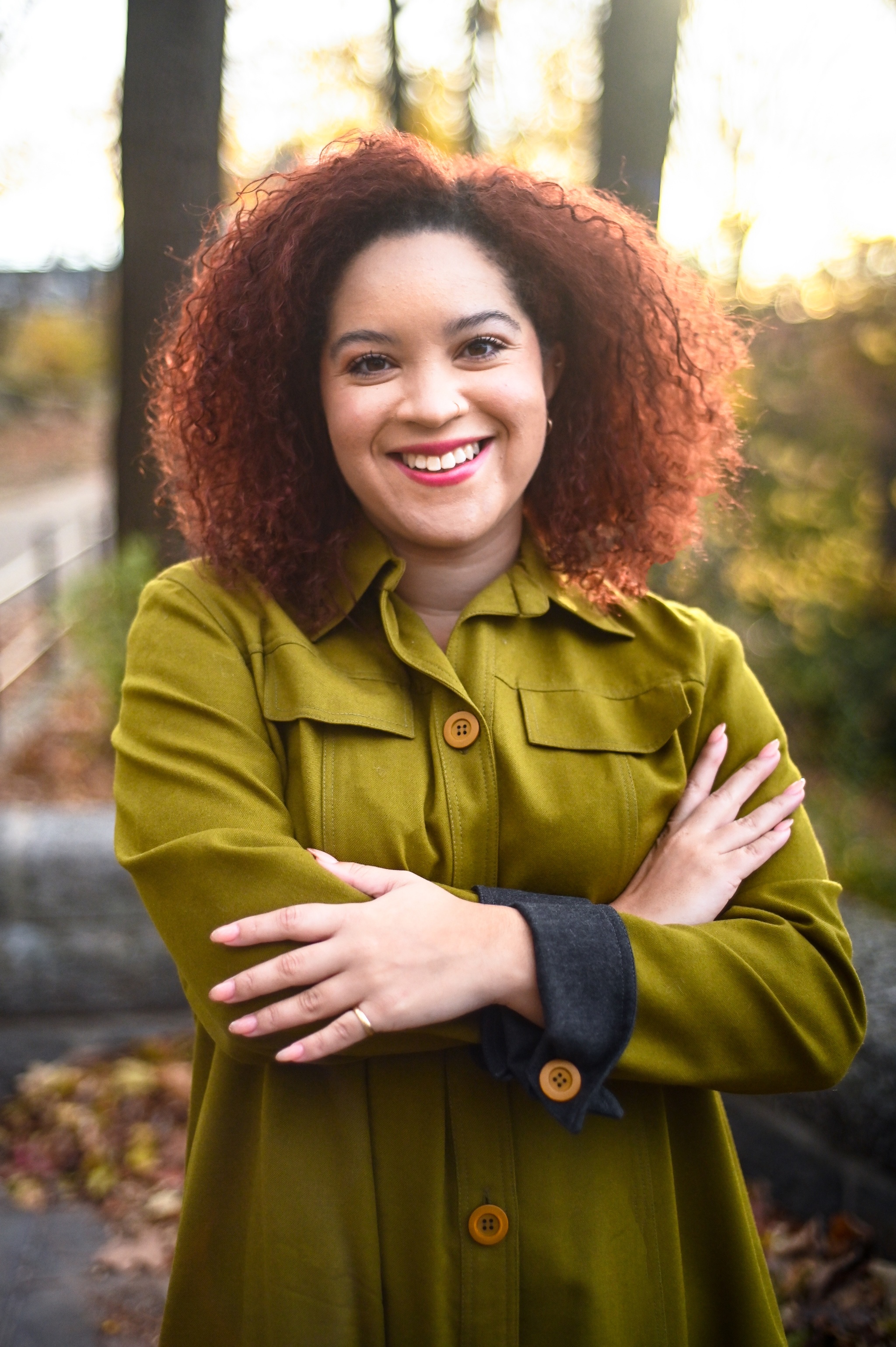 A portrait of Adeze Wilford, a Black woman with light skin and auburn hair who poses smiling with her arms crossed across her chest. Adeze is outdoors with trees and foliage behind her. She wears a green dress that fades from lighter to darker green.
