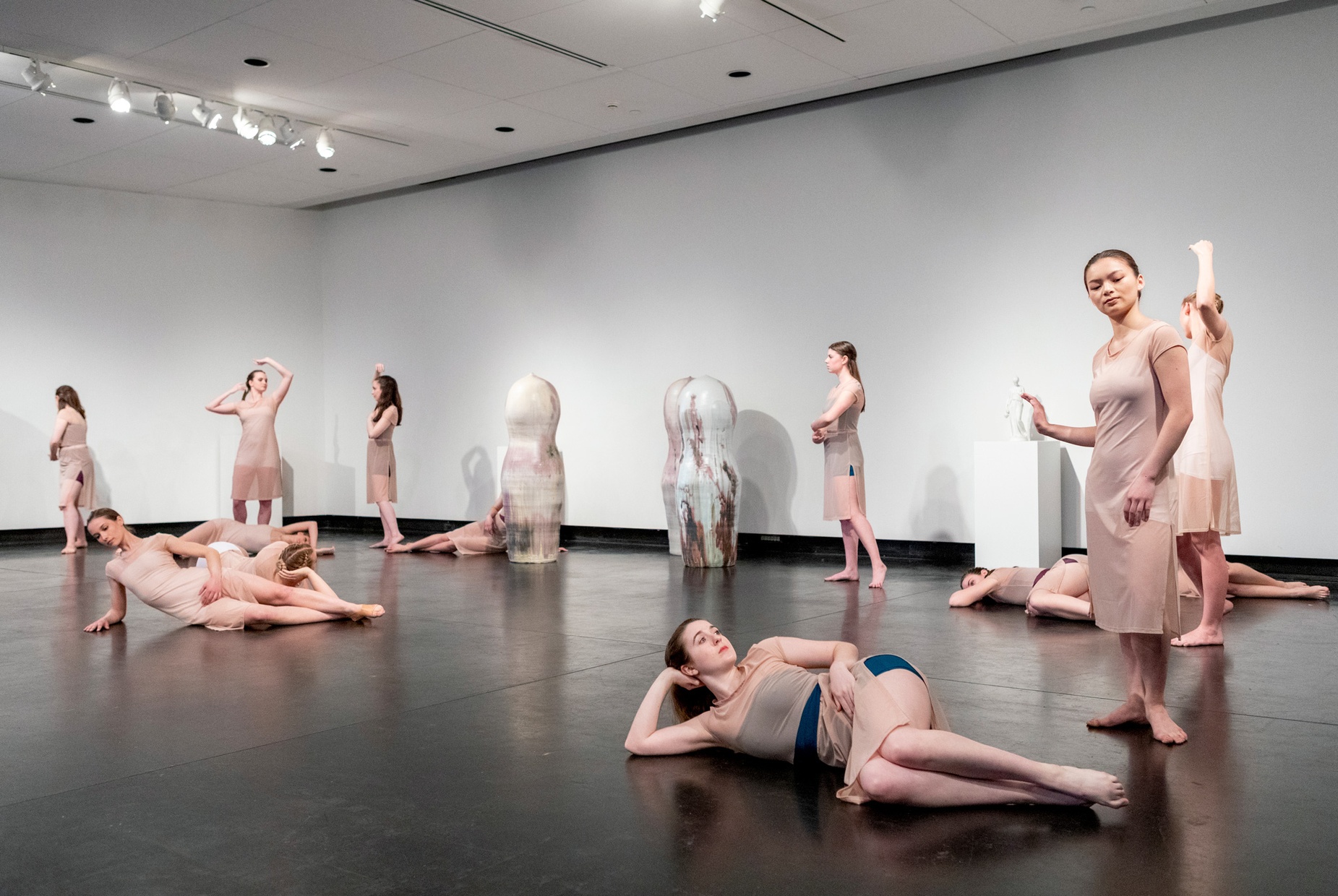 A group of female dancers in nude dresses pose in a gallery space around two, white oblong sculptures.
