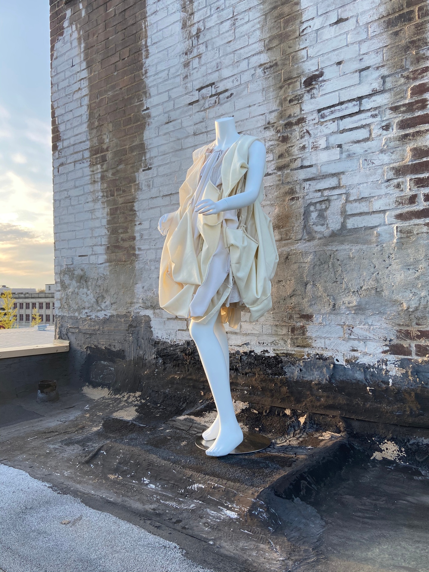 Mannequin wearing an off-white shift dress made of bunched and draped fabric. Mannequin is placed on a rooftop against a dirty whitewashed brick wall.
