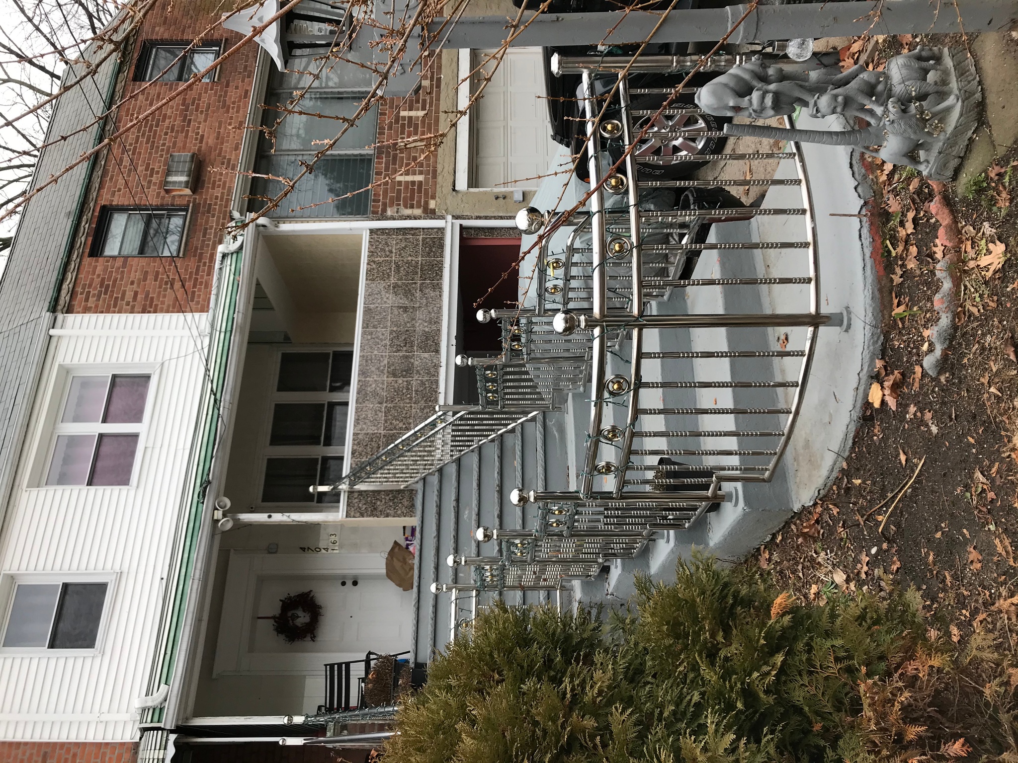 A white clapboard row house with a long gray concrete stairway leading to the front door. A shiny steel railing lines either side of the stairs, curving as the stairs join a short sidewalk that leads to a driveway