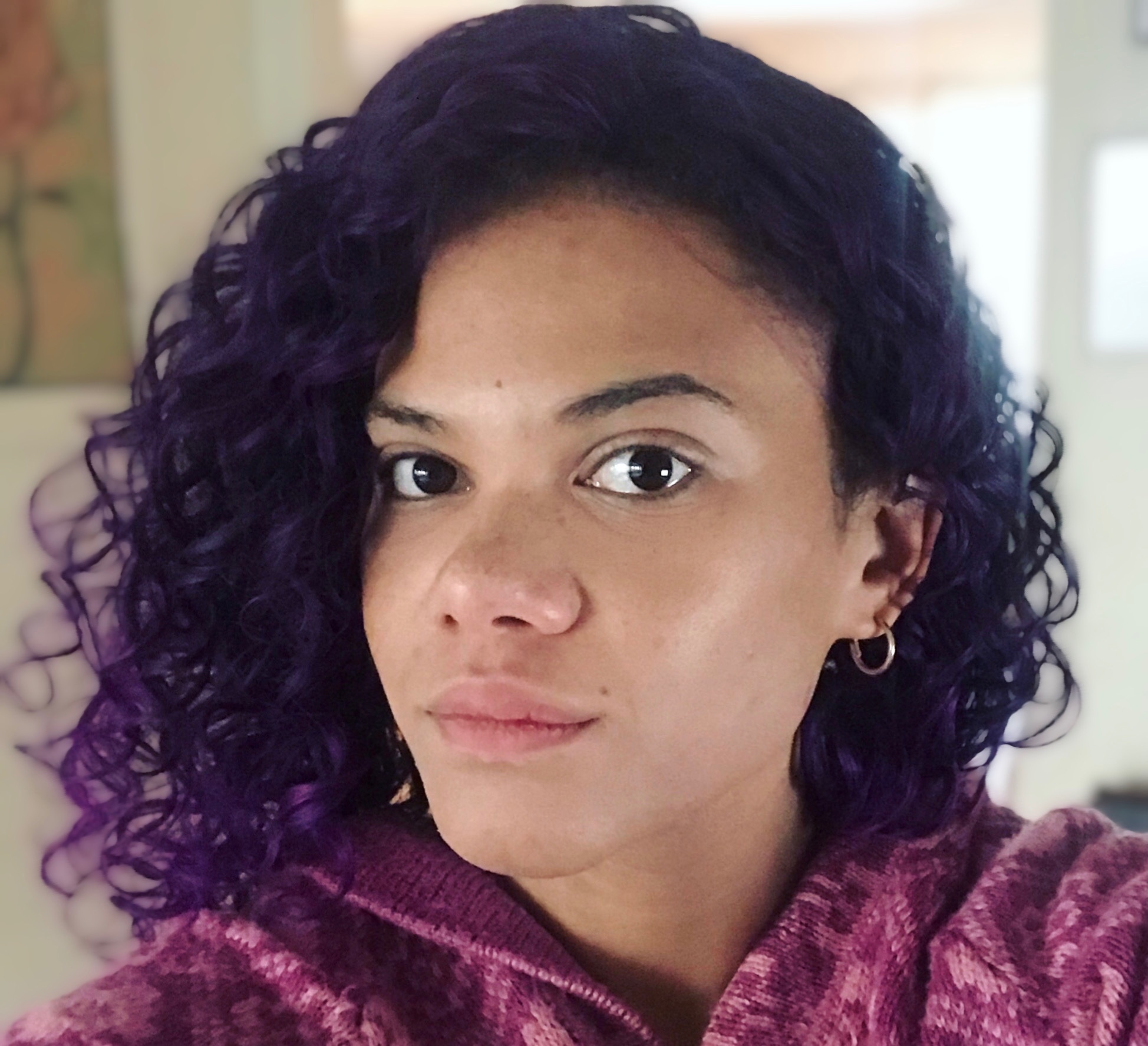 A woman with curly purple hair parted to her right side. She looks at the camera as if holding it herself for a selfie. 