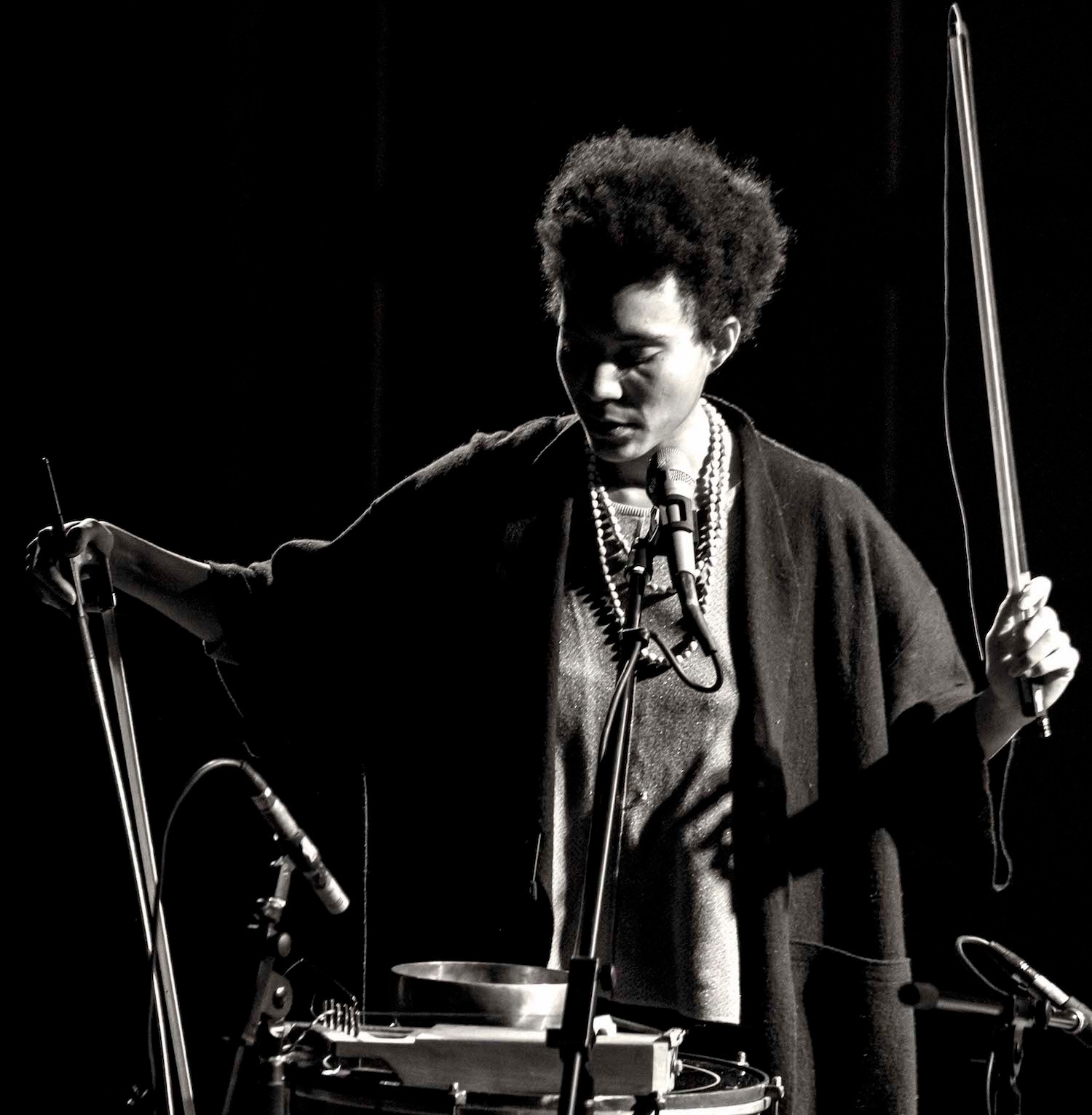 A woman holding two string instrument bows, one pointed up and one pointed down, as she stands playing instruments in front of a set of microphones