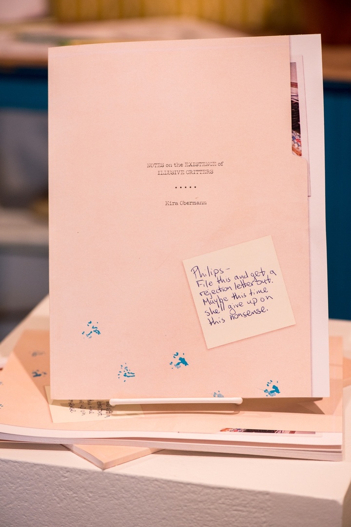 Manilla folder stamped with "Notes on the Existence of Elusive Critters - Kira Oberman." A sticky note on the file reads, "Philips - File this and get a rejection letter out. Maybe this time she'll give up on this nonsense."