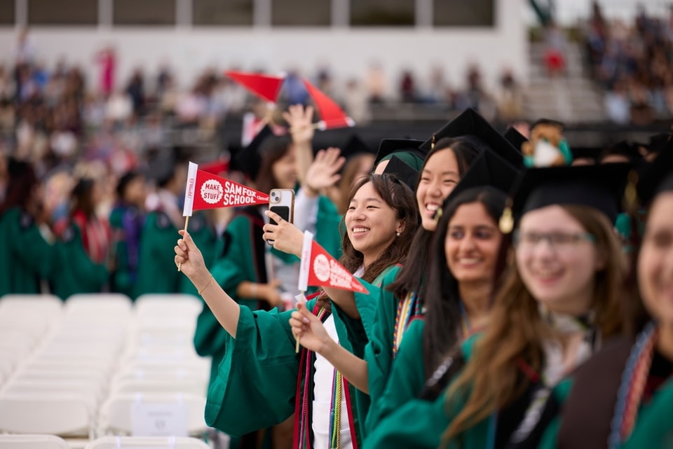 A group of students sit in a line while wearing regalia and carrying pennants for the Sam Fox School.