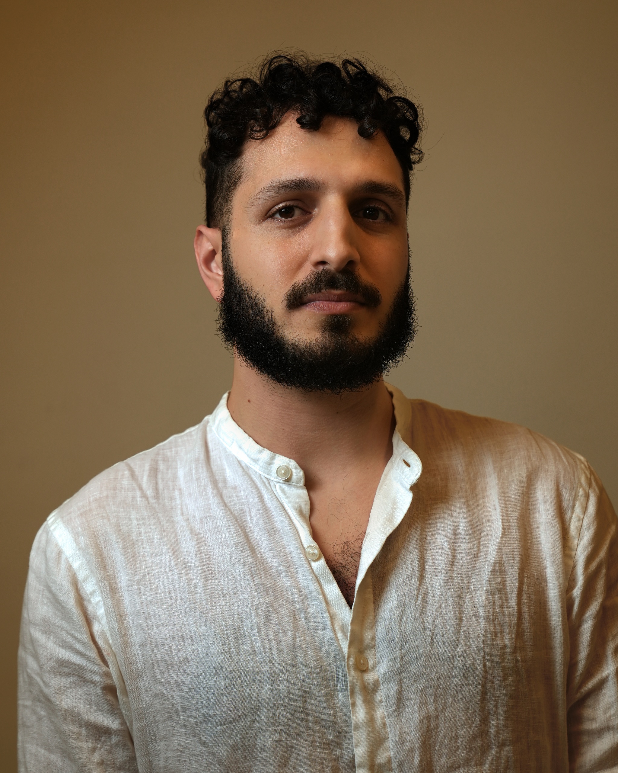 A portrait of Waseem Alzer against a neutral brown background. Waseem wears a white shirt with the top buttons undone. He has short curly hair, a dark beard, and looks directly at us. 
