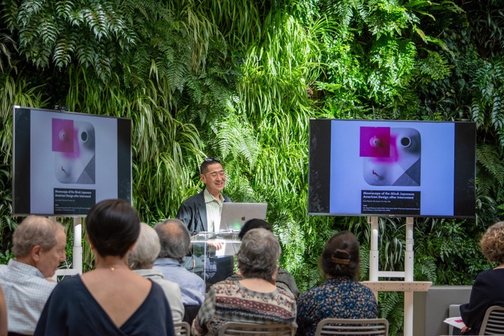 Oshima stands at a podium in front of two screens displaying an image of a square white sculptural tile with a pink plexi overlay. The slide reads "Moonscape of the Mind: Japanese American Design after Internment." Oshima smiles and addresses a seated audience.