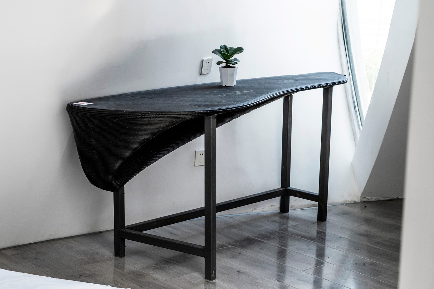 A black, 3-D printed side table with four legs. The top of the table is curvilenear, and skinnier at the far end. Toward the front of the table, the surface has depth underneath.