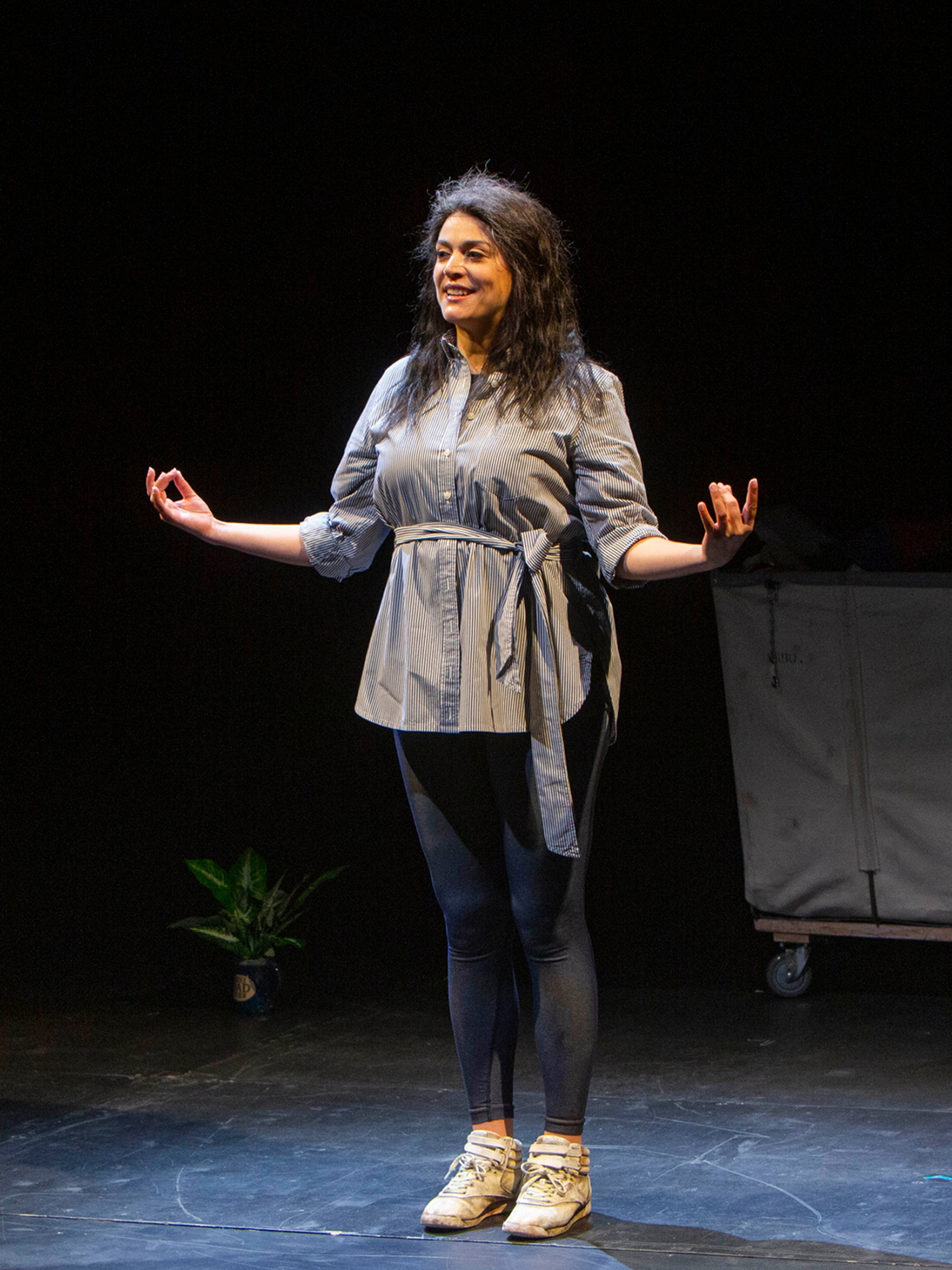 The performer Cecily Strong on a black box theater stage wearing a wig with graying hair and holding her arms outstretched