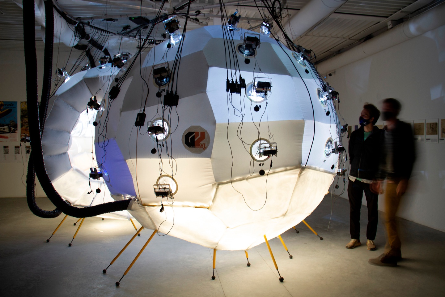 A split-sphere, multimedia installation in a darkened room, with two people looking at it. The sculptures are two large, seemingly inflatable paneled spheres, tied down with cords and lit from underneath, with rounded portholes for light and cameras.