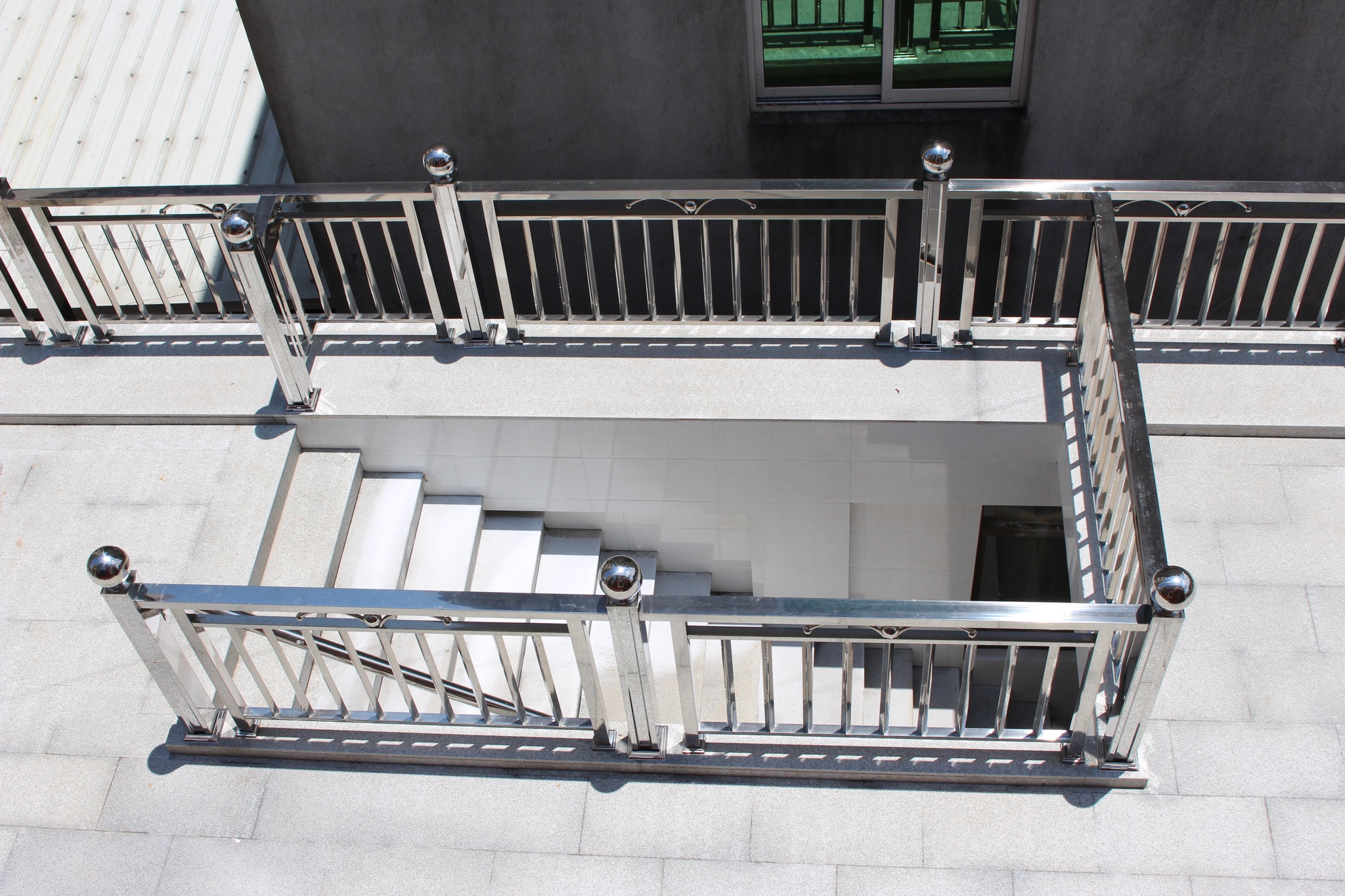 An aerial view taken from above of a set of stairs descending into a concrete patio, surrounded by a gleaming steel banister and railing. A neighboring house faces the banister at the top of the image, with a green-glass window reflecting some of the banister.