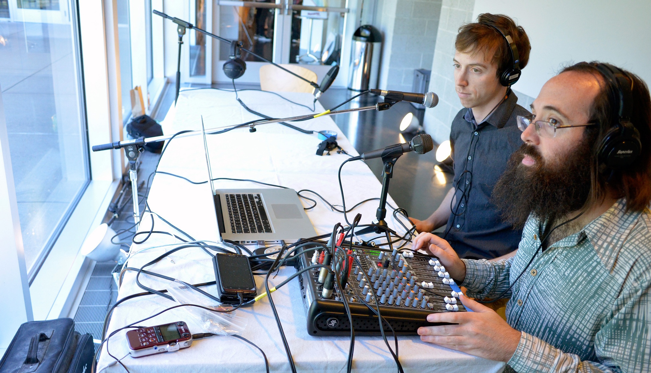 Two white men, one older and one younger, sit at a white table with mic stands and a sound board facing a wall of windows.