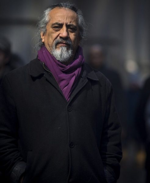 A portrait of george emilio sanchez. He stands outside in what seems to be a city environment wearing a purple scarf around his neck and tucked into a dark colored overcoat. He has gray hair and a gray mustache and goatee.