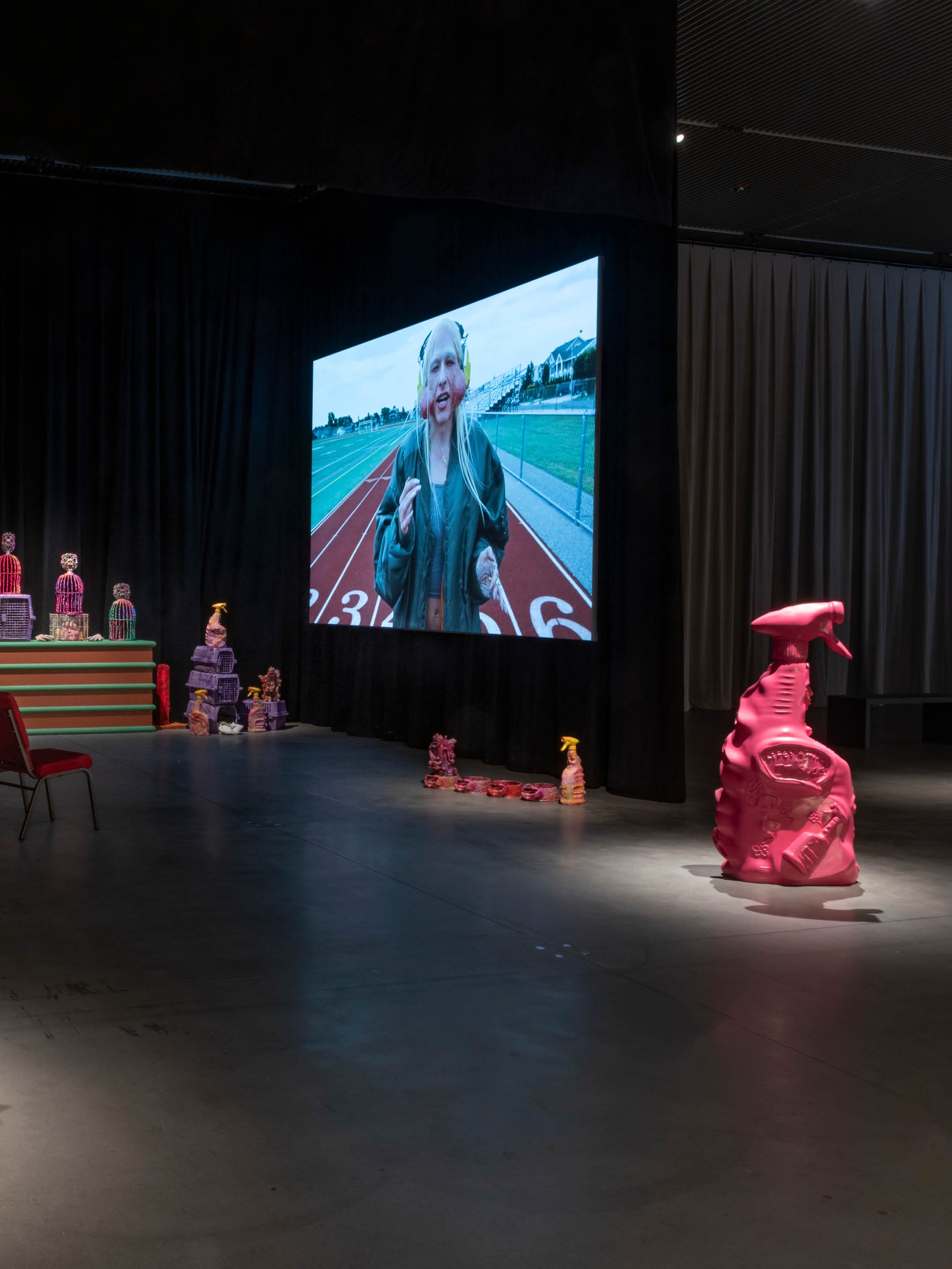 An installation of sculptural objects and a film in a gallery with low light. The sculptural objects are oversized versions of pet products, like cages, dog bowls, and cleaning supplies. They are cast in fluorescent colors. In the foreground is a large pink spray bottle sculpture, about four feet tall. In the background, the film projected on a screen shows the artist Jake Brush in character with a long blond wig and prosthetic jowls protruding from his cheeks while he walks on a high school track.