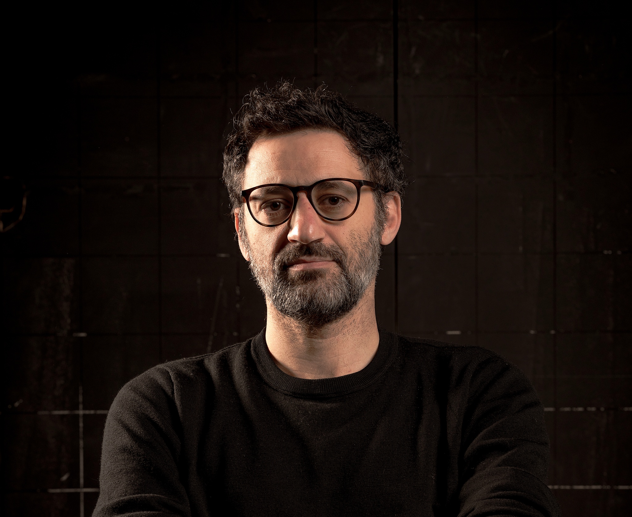 A portrait of director Omar Elerian. He is seen from the shoulders up against a dark background. He wears a dark sweater that blends in to the background. He has a scruffy, short beard, short dark hair, and wears glasses.