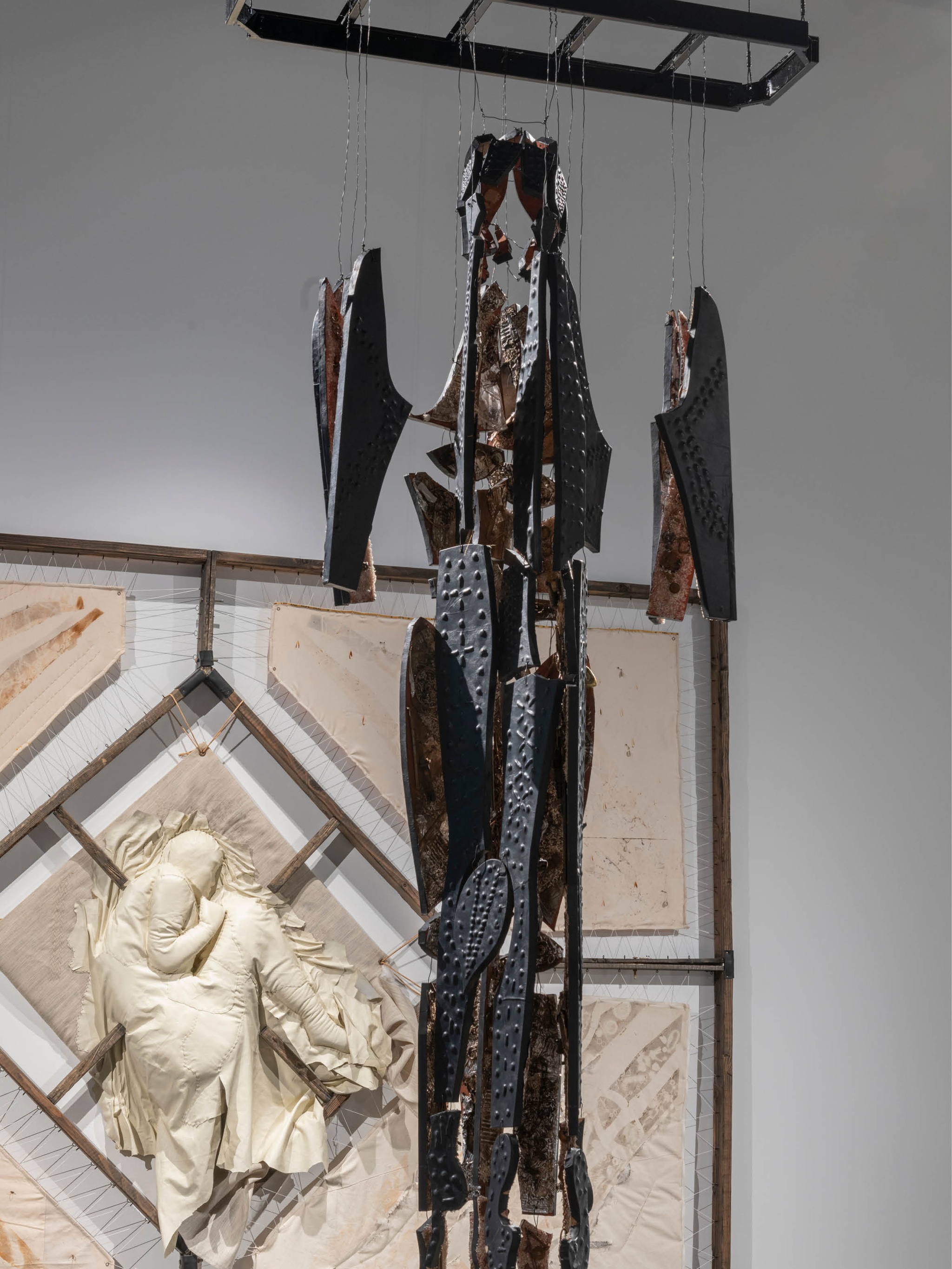 An installation of five artworks in a gallery. They are placed on the ground, hanging from a wall in the background, or hanging from the ceiling. The works on the ground include a box with leather scraps piled inside, a white piece of leather dropped on the floor, and a clothing rack similar to a coat rack. On the wall, a tapestry is stretched on a metal frame. At its center a human figure is formed out of white leather that has been stuffed. The figure is in the fetal position. Hanging at the center of these works is a pattern of the artist's body that has been cut out of black leather and affixed to shaped wood panels. The individual parts are attached by wires similar to a skeleton. 