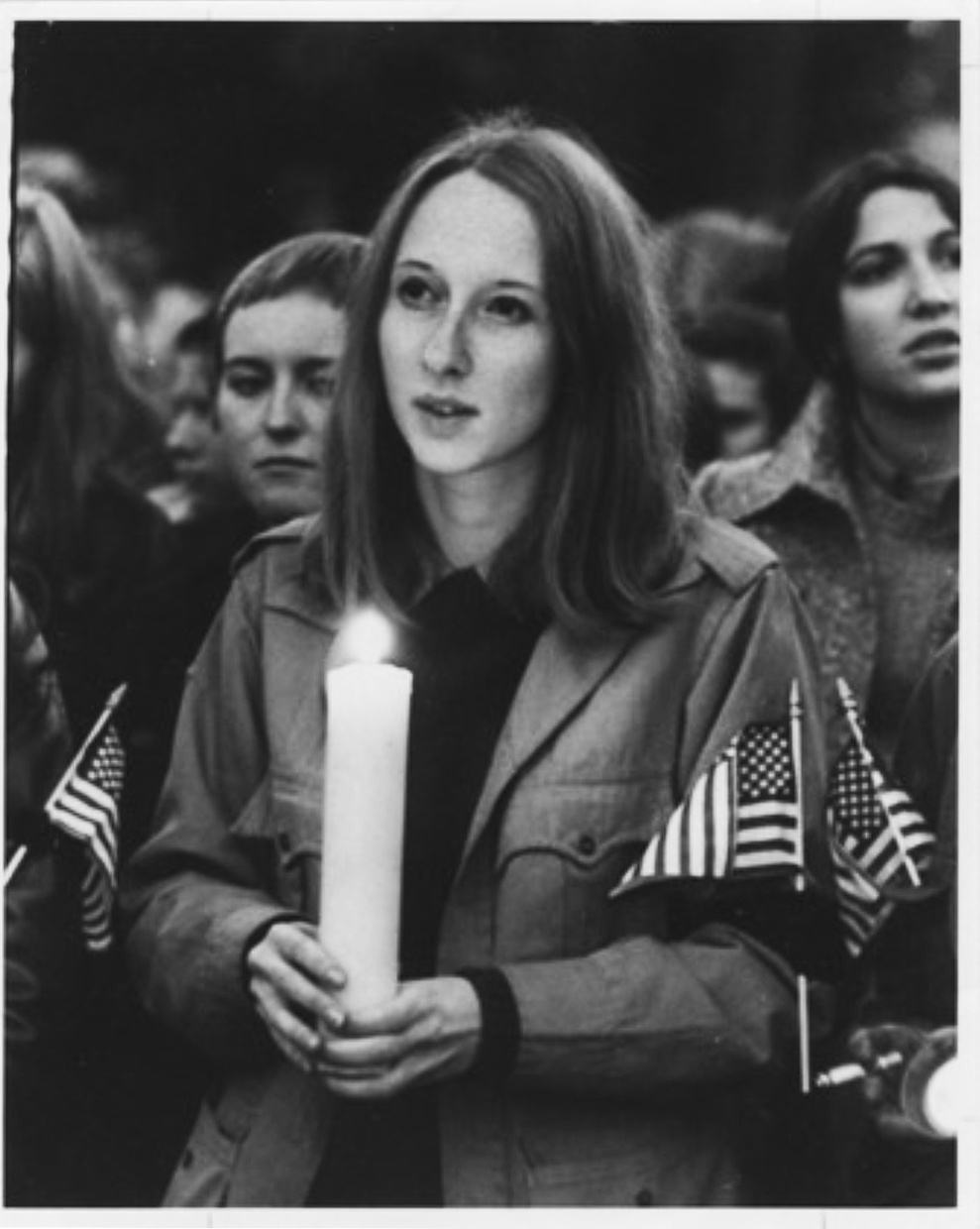 A slightly blurred black and white image depicts a young white woman with dark shoulder length hair holding a large, lighted candle, wearing a coat with the American flag attached to one of her sleeves.
