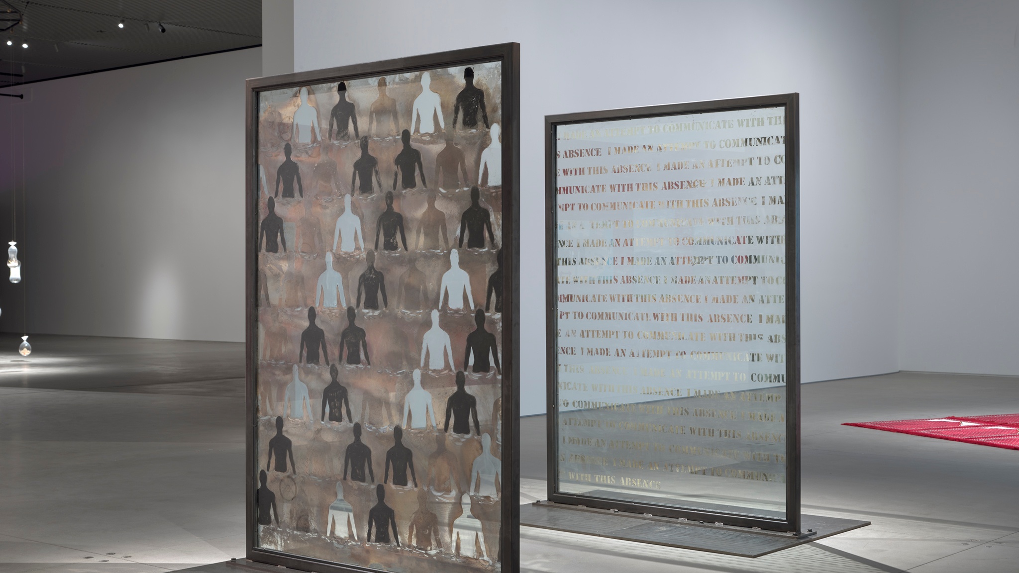 Two glass panels standing upright in a gallery. One has the form of a person from the waist up stenciled in a repeating pattern of mirrored, painted, and transparent glass. The other repeats the sentence "I made an attempt to communicate with this absence.”