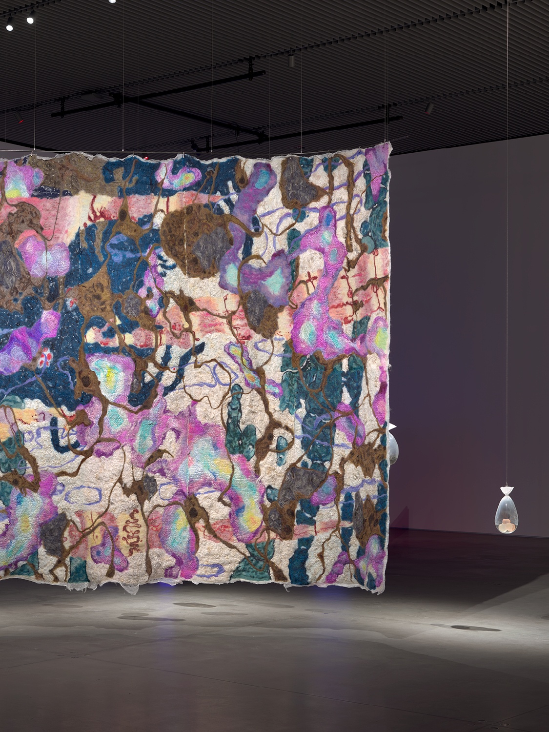 An abstract felt tapestry made of squiggly lines and shapes of predominantly purple and pink tones. To the side, a glass object hangs to counterbalance the tapestry.