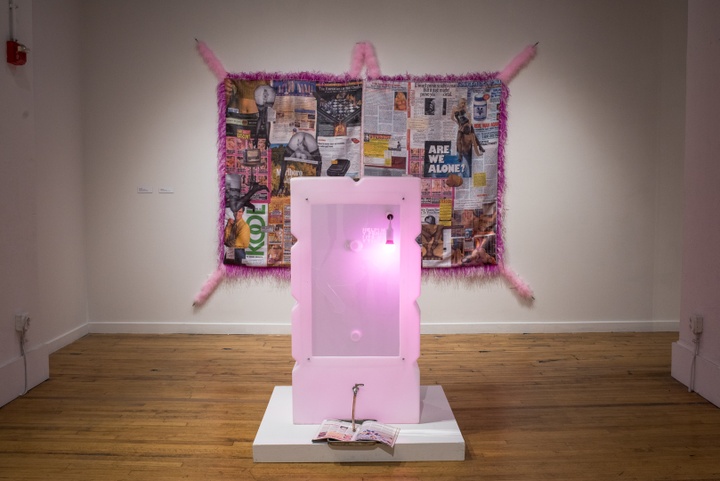 A pink glowing box the size of a minifridge with various nozzles and notches sits on a small dais on the floor. Hung on the wall behind it is a quilt of sensationalist magazine clippings bordered by a pink feather boa.