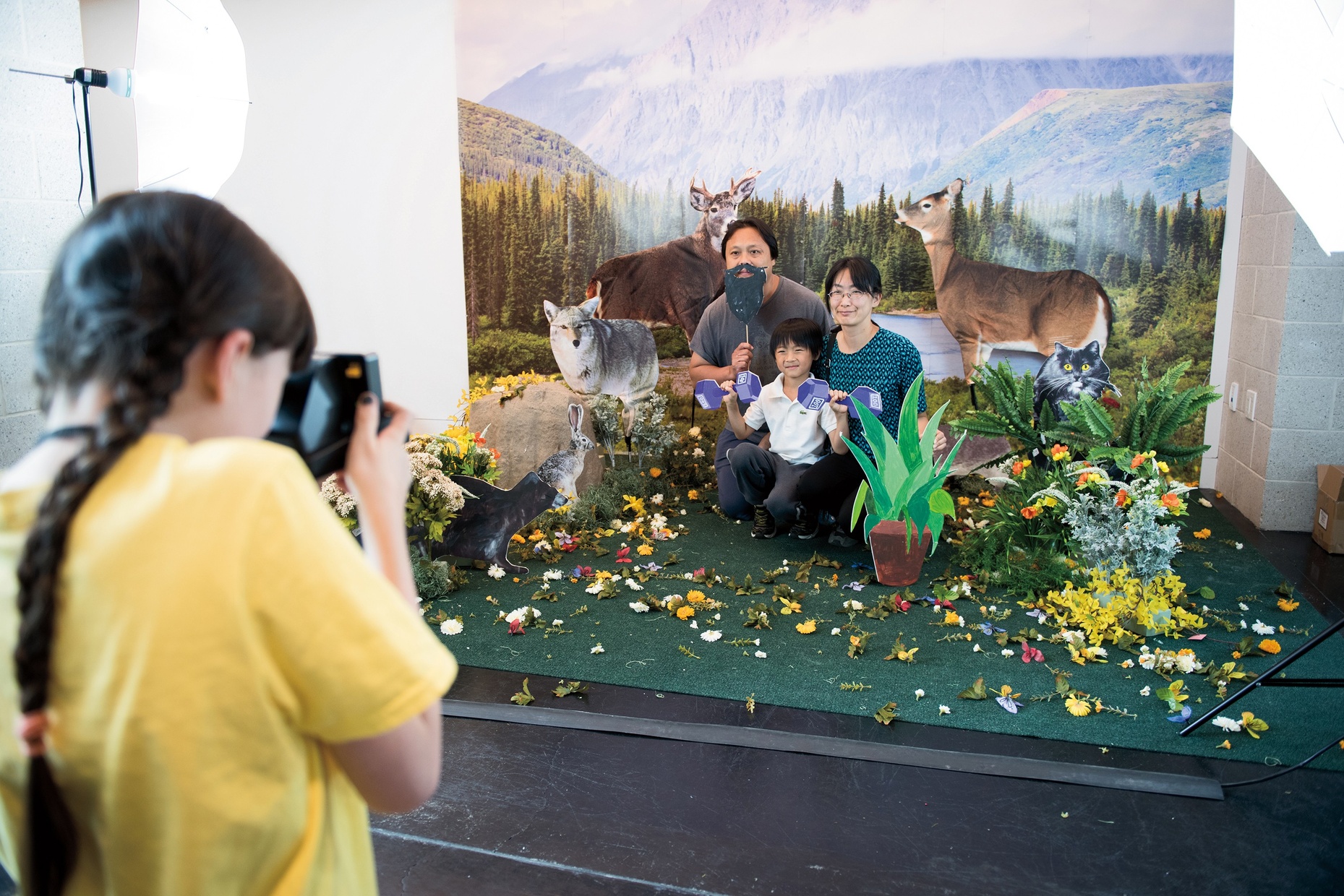 A young, light-skinned girl takes a Polaroid picture of a small, light-skinned family smiling and posing in a fake, nature diorama.