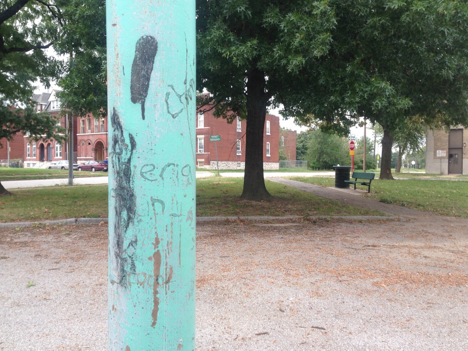 Close-up view of a weathered teal green pole, with pavement and trees behind it.